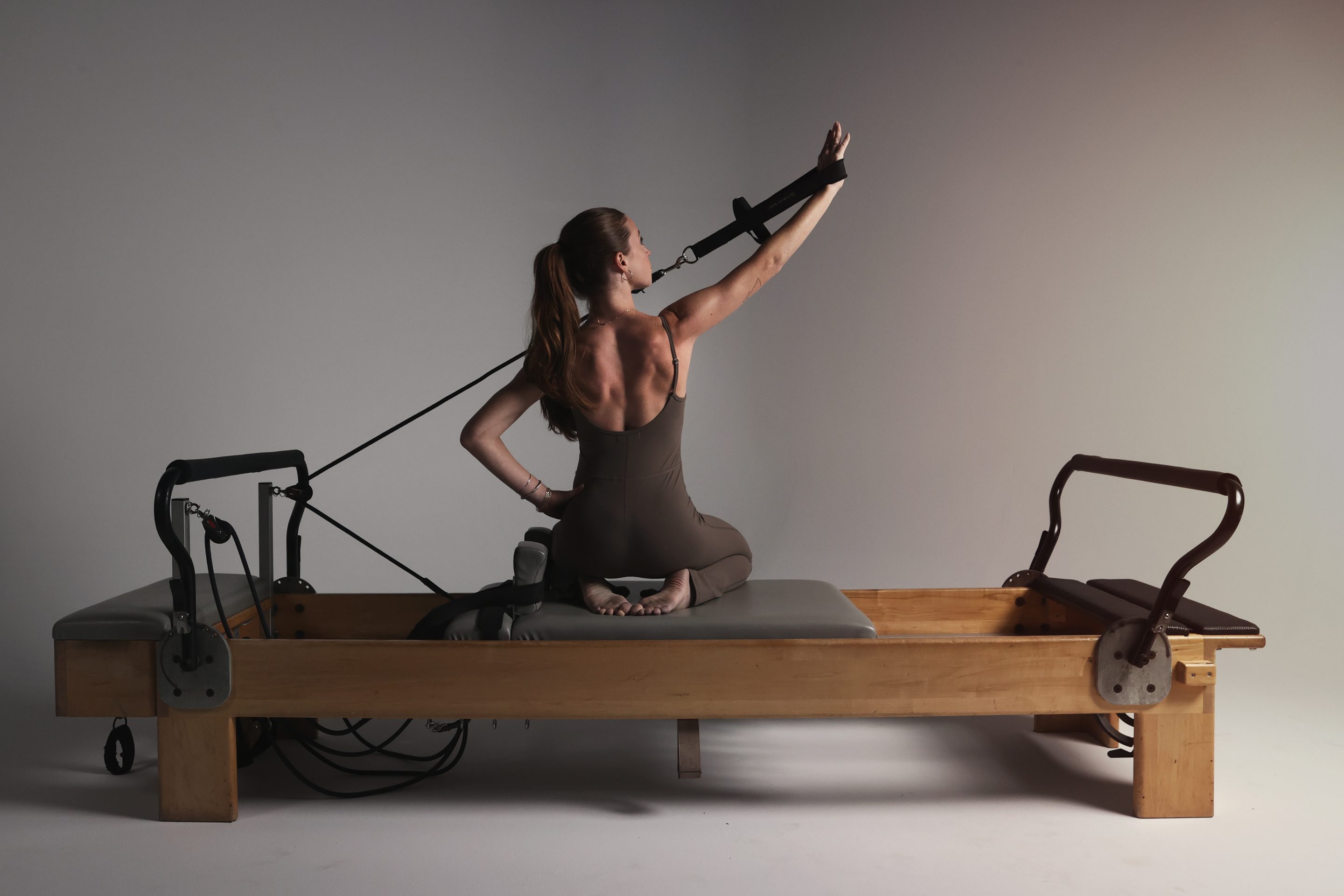 A woman performing a Pilates exercise on a wooden reformer machine, kneeling with one arm extended and resistance straps engaged in a minimalist studio setting.