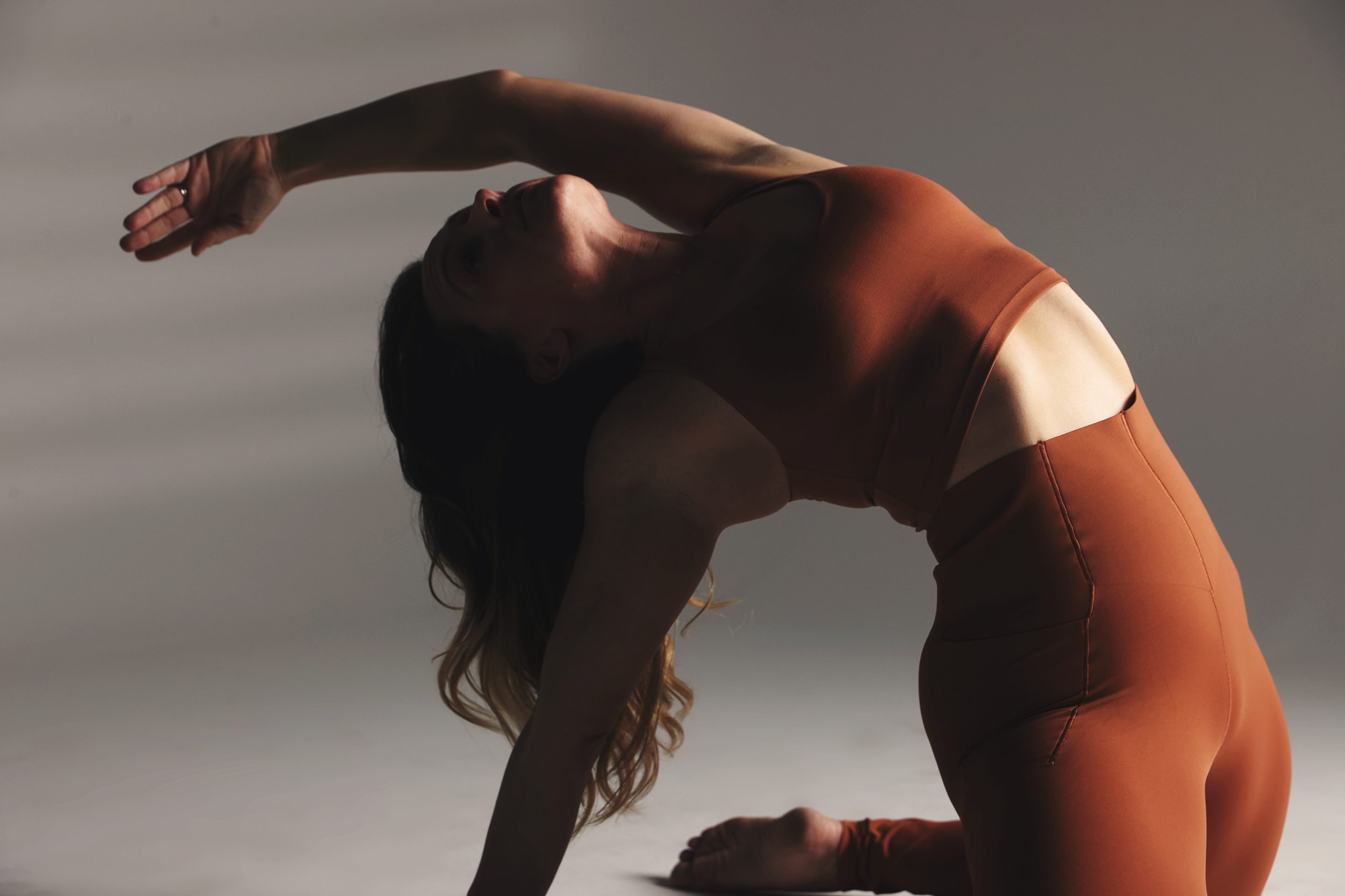 A woman in a rust-colored outfit practicing a graceful yoga backbend in soft studio light.